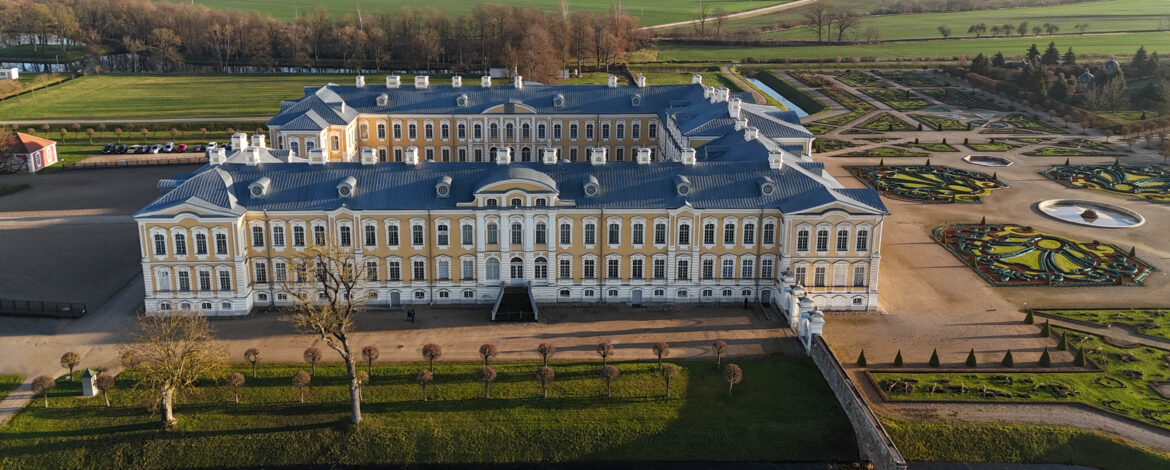 Attēlā redzama Rundāles pils Latvijā – iespaidīga baroka un rokoko stila celtne ar dzeltenām fasādēm un baltām detaļām. Attēla priekšplānā redzams kopts zāliens ar kanālu un simetriski stādīti koki, kas apcirpti lodes jeb apaļā formā. Pils labajā pusē daļēji redzams franču stila dārzs ar ģeometriskās figūrās veidotu parteri un strūklaku partera centrā. Tālāk aiz pils fonā atrodas plaša, lauku ainava ar kokiem un pļavām.