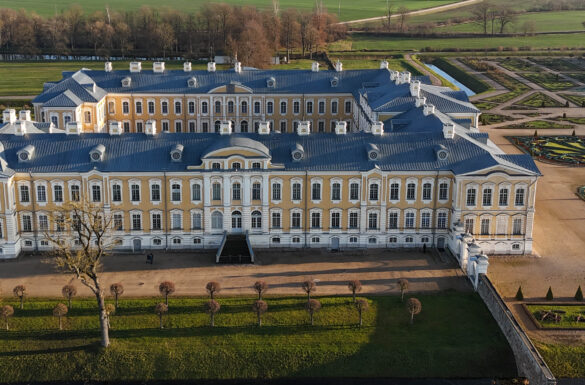 Attēlā redzama Rundāles pils Latvijā – iespaidīga baroka un rokoko stila celtne ar dzeltenām fasādēm un baltām detaļām. Attēla priekšplānā redzams kopts zāliens ar kanālu un simetriski stādīti koki, kas apcirpti lodes jeb apaļā formā. Pils labajā pusē daļēji redzams franču stila dārzs ar ģeometriskās figūrās veidotu parteri un strūklaku partera centrā. Tālāk aiz pils fonā atrodas plaša, lauku ainava ar kokiem un pļavām.